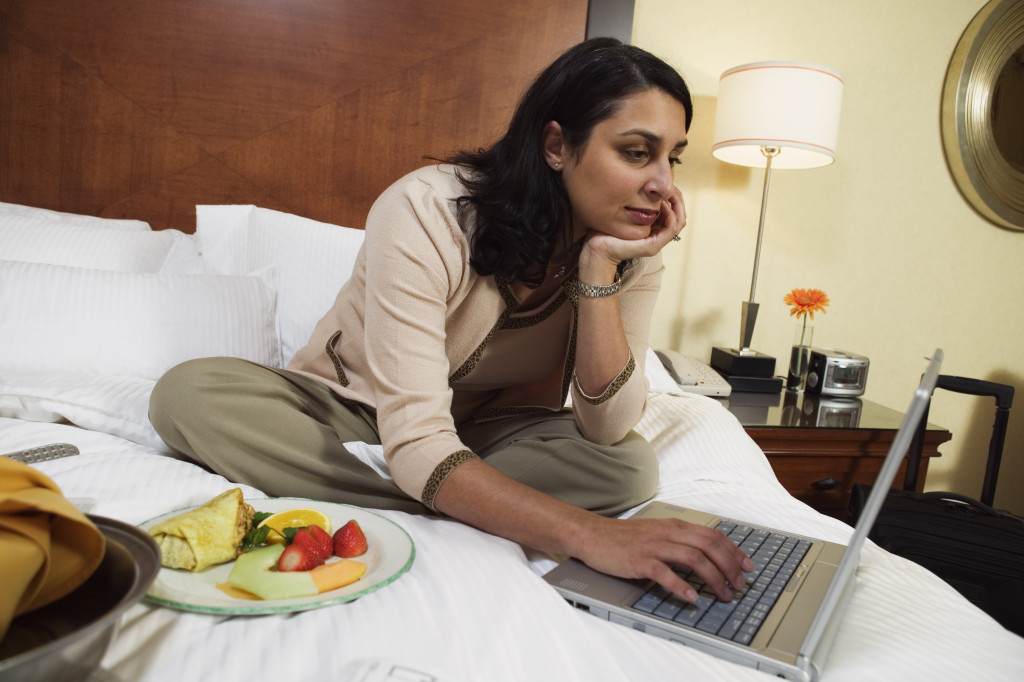 Businesswoman working on laptop and eating food on bed in hotel room