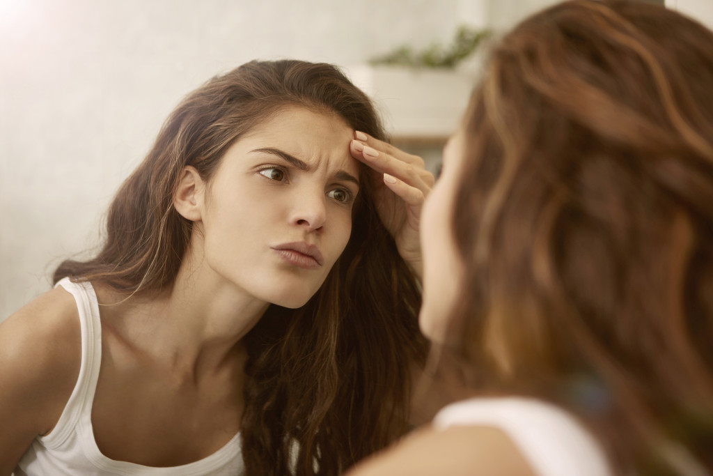 A woman looks closely at a pimple on her forehead in a mirror